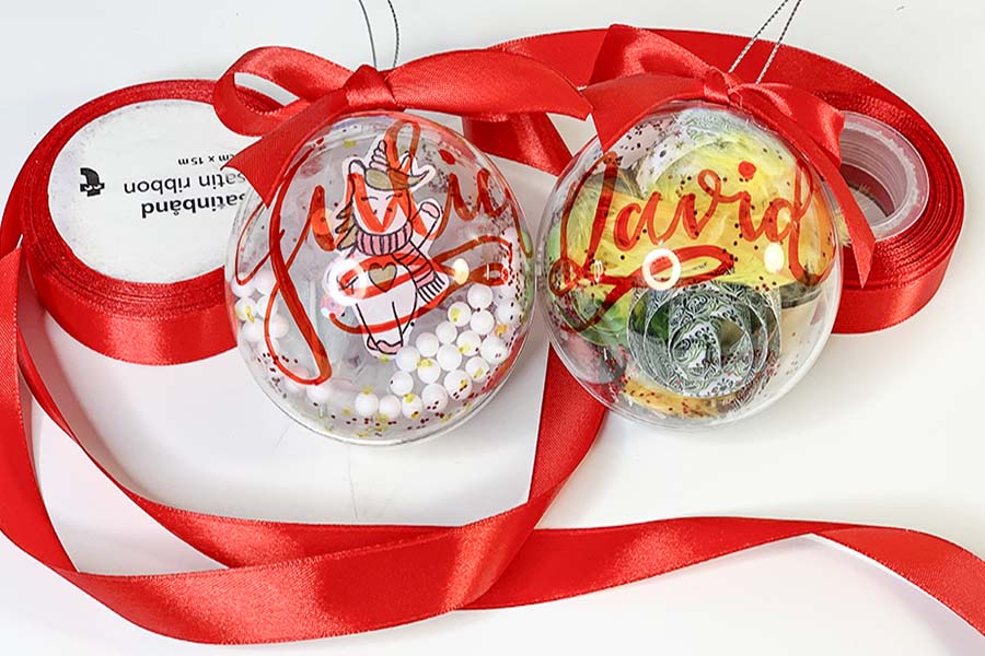 dos bolas de navidas transparentes en las que hay escritos dos nombres y alrededor de ellas un cinta roja