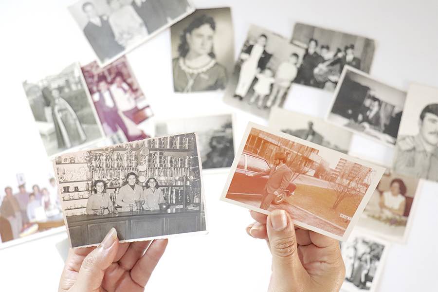 en primer plano dos manos sostienen fotos antiguas y al fondo sobre una mesa varias fotos puestas de manera aleatoria